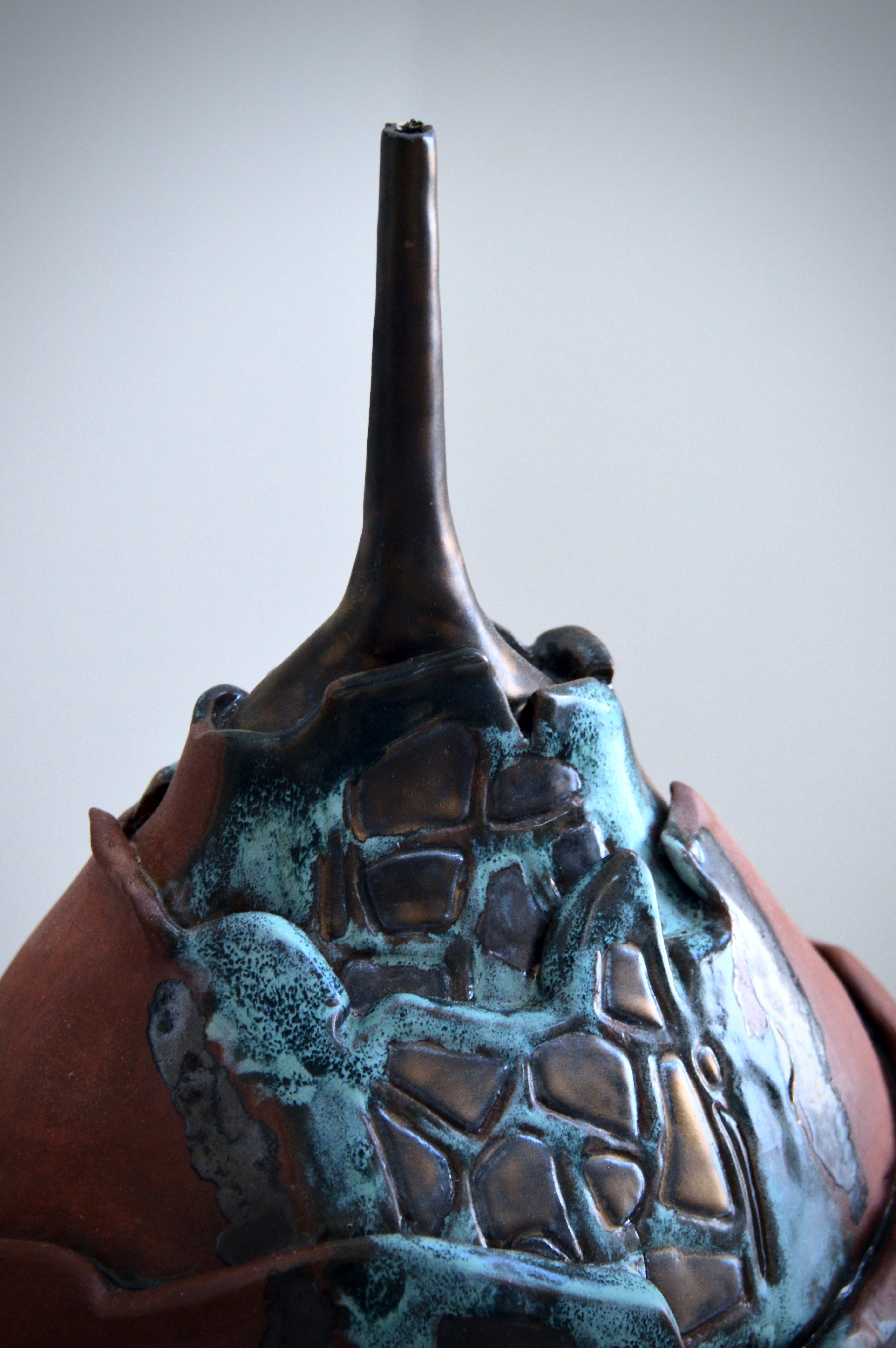 Close-up of a textured, turquoise and copper ceramic vase with a long neck on a light gray background