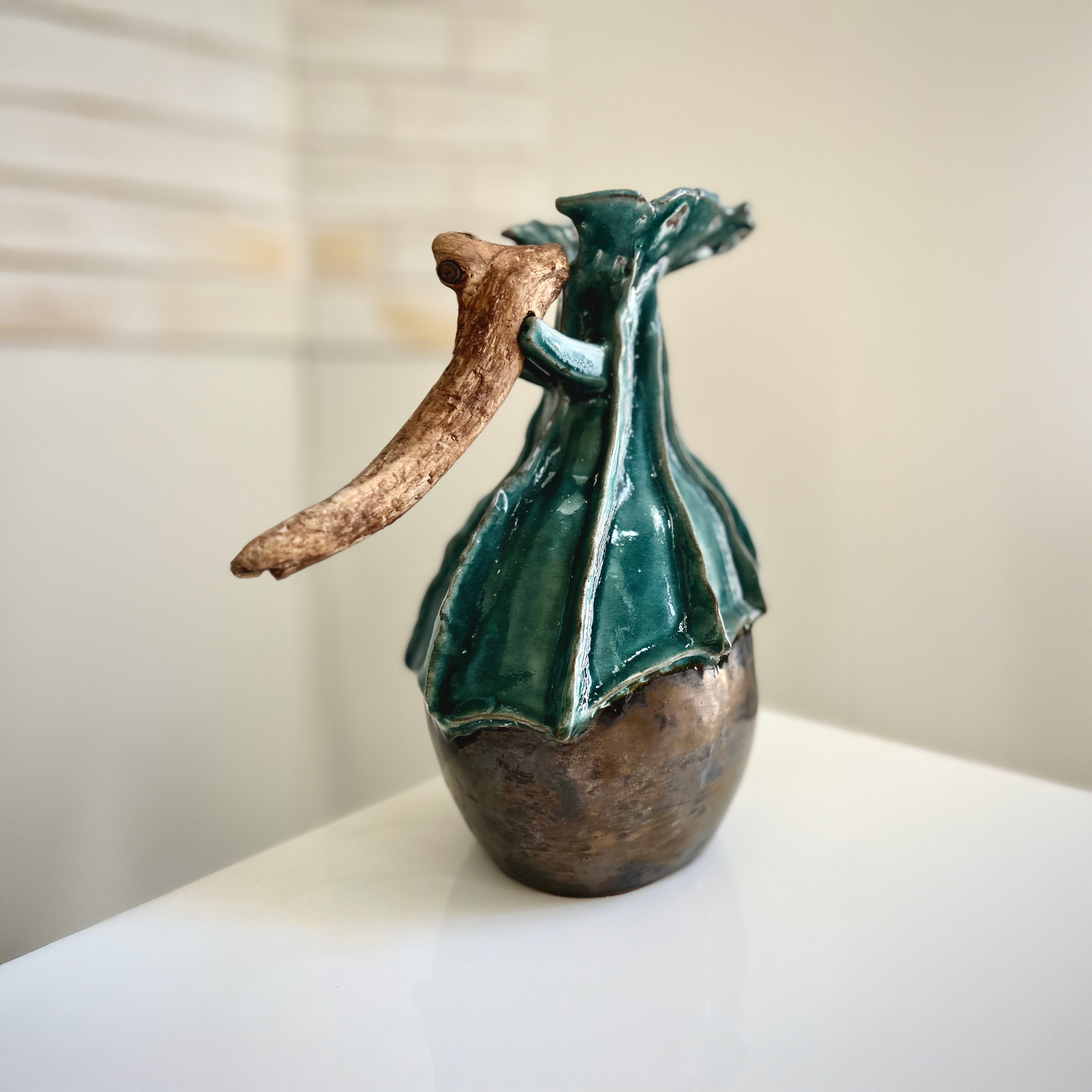 Ceramic vase with green glaze and brown accents on a white surface with a wooden handle