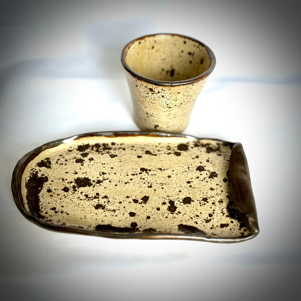 Speckled ceramic cup and rectangular plate on a reflective surface with a blurred background